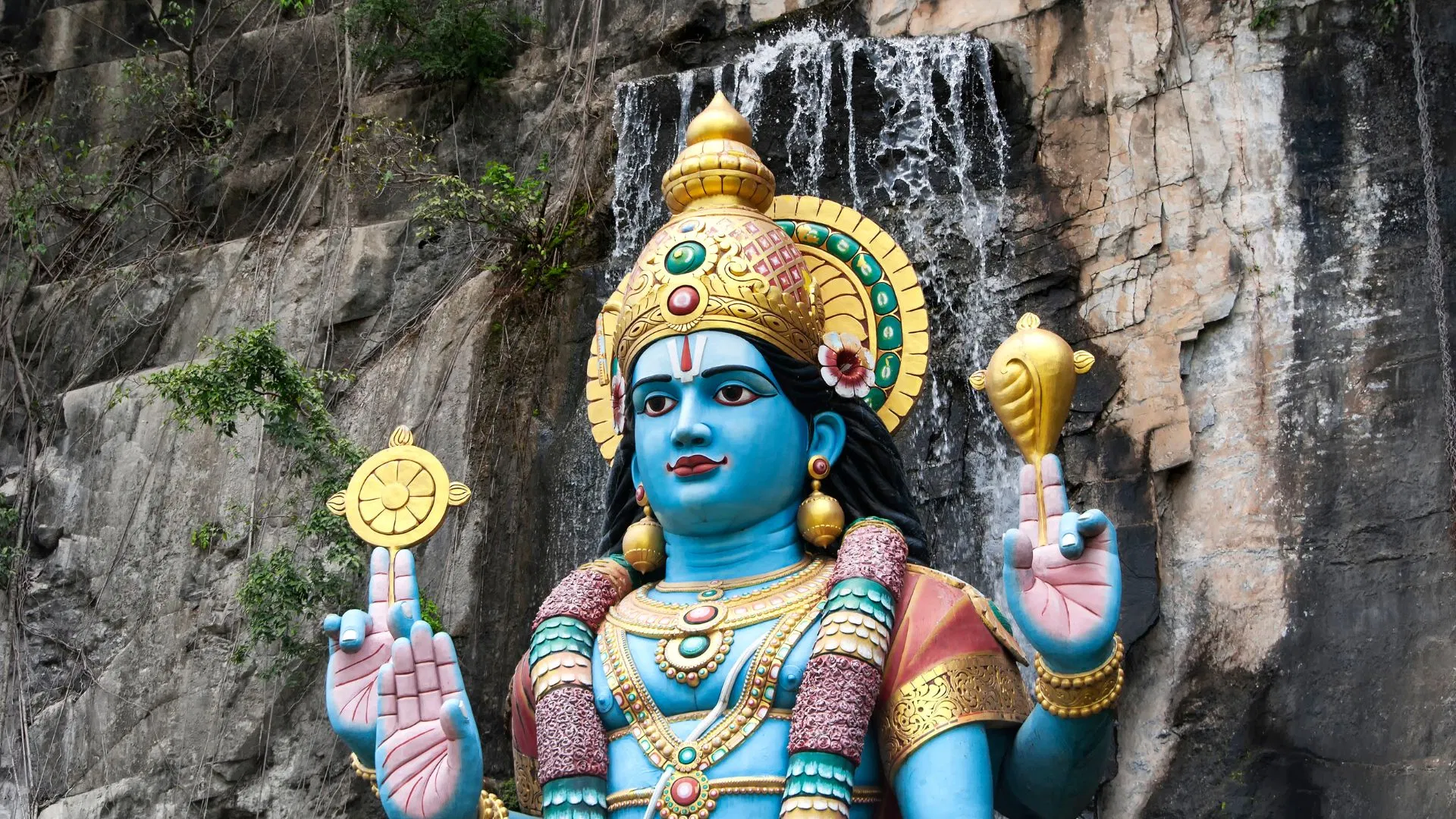 A large blue four-armed statue standing below the limestone cliff at Batu Caves, with water running down the rock face